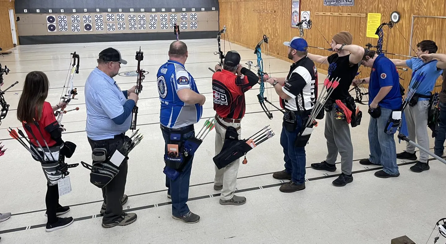 view from behind a line of adult archers on the firing line of the indoor range. In front of them is a wall covered in paper targets