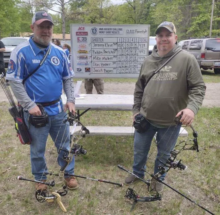 Two adult archers with their bows in front of a score board outdoors