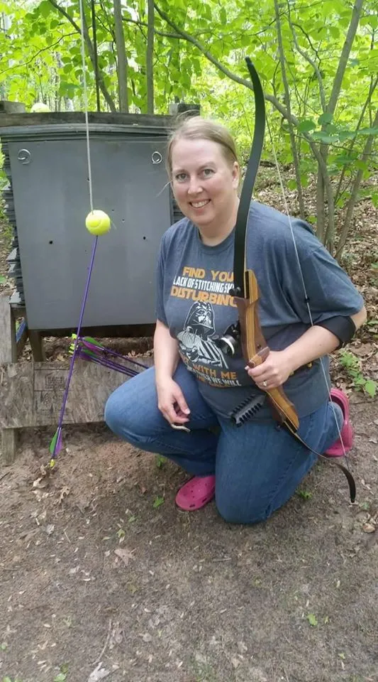 A smiling women holding a recurve bow next to a tennis ball suspended on a string with an arrow in it