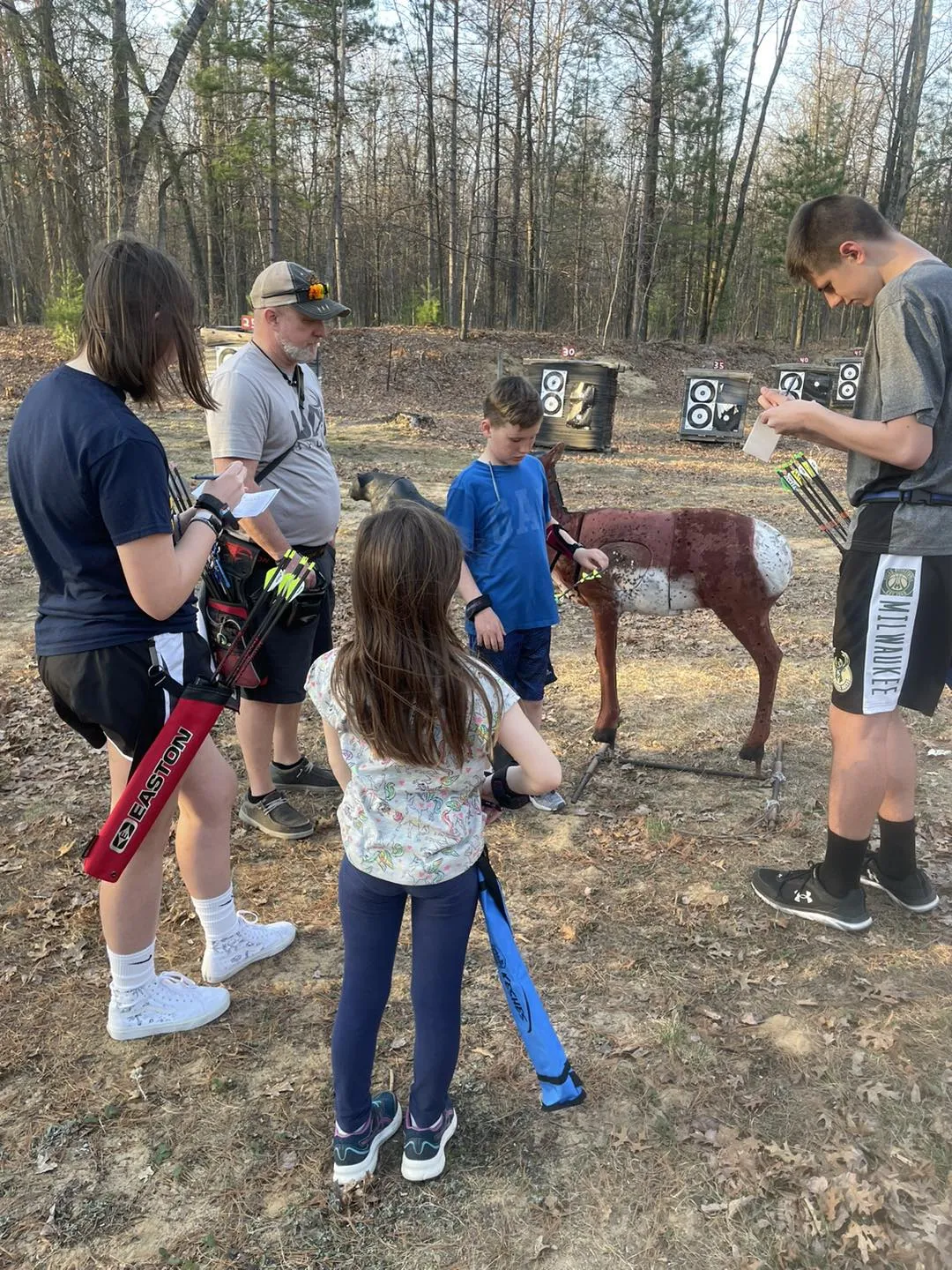 One coach and 4 youth archers standing near a 3D deer target with arrows in it. The older youth archers are recording scores on score cards