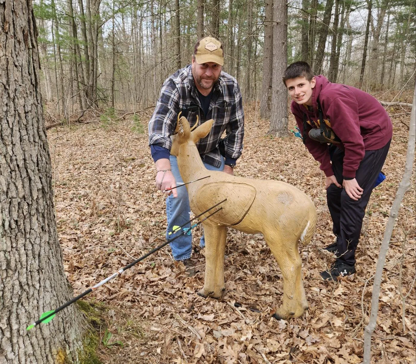 A coach and youth archer standing behind a 3d target with several arrows in it. The coach is proudly pointing out the archer's arrow that has perfectly hit one of the existing arrows, known as a robin hood